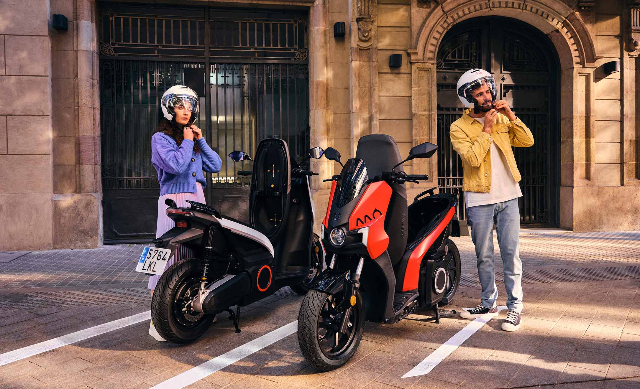 Elscooter med två säten sedd från sidan. Den som är sedd framifrån är röd och har seat mo-logotypen. Den andra är mörk och grå och parkerad bakifrån från sidan. De omges av ett stadslandskap och åtföljs av en individ som tar på sig hjälmen.