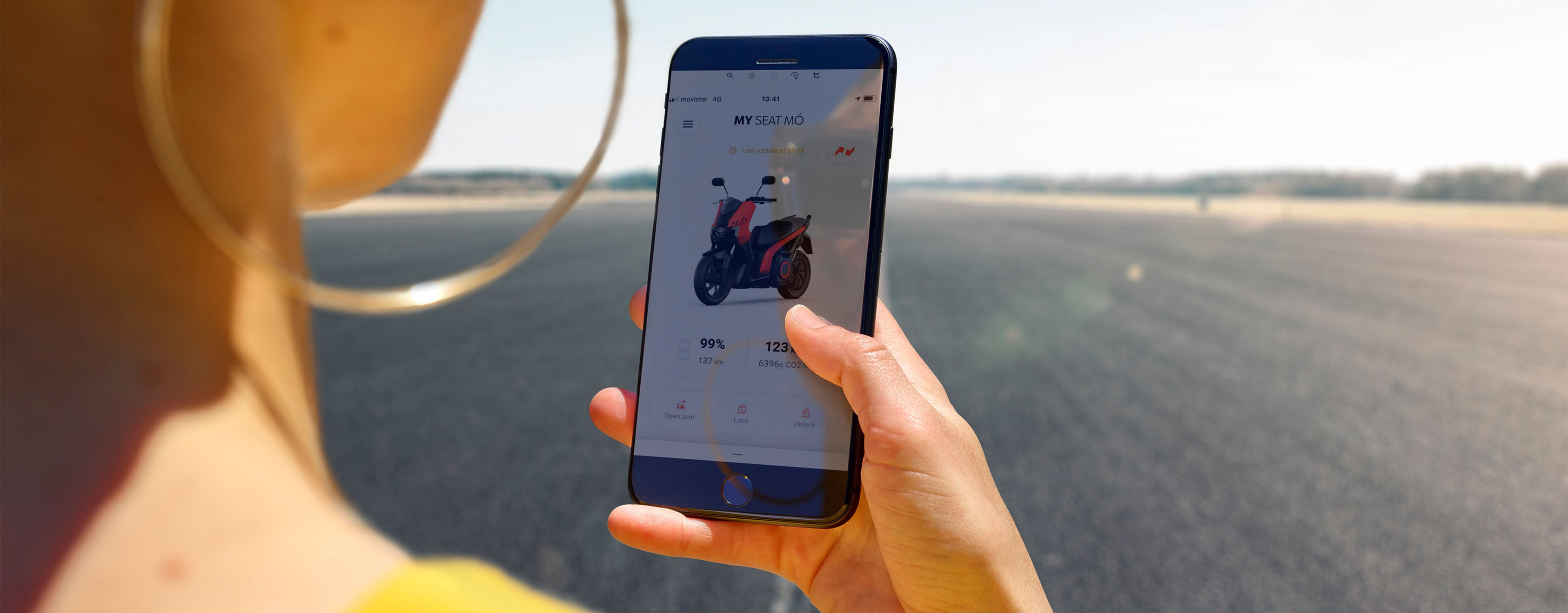 En person som håller en smartphone och använder my seat mó-app i en avlägsen solig miljö, och som betonar tanken på att kunna komma åt din scooters funktioner när som helst, var som helst.