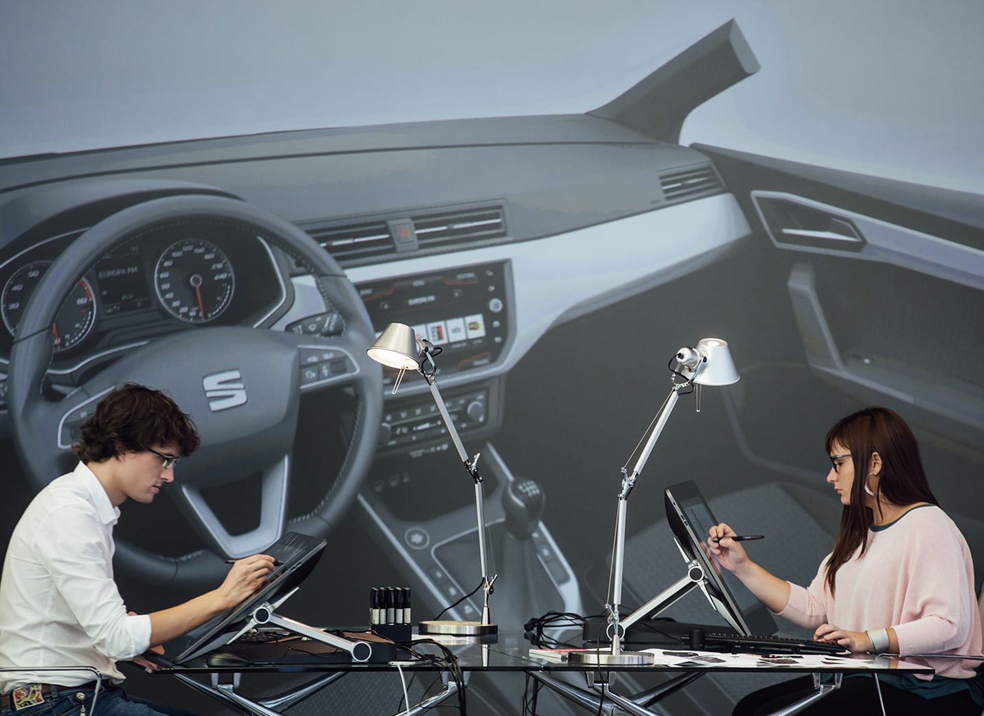Två nybilsdesigners ritar på elektroniska indikeringsenheter mot en fotobakgrund som visar insidan av en bil – SEAT Human Resources