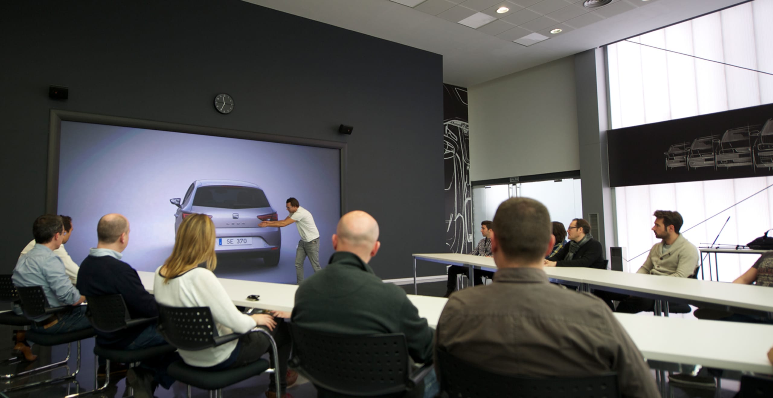En man gestikulerar mot en SEAT Leon-bildesign vy bakifrån med människor som sitter vid ett bord och tittar på – SEAT Human Resources