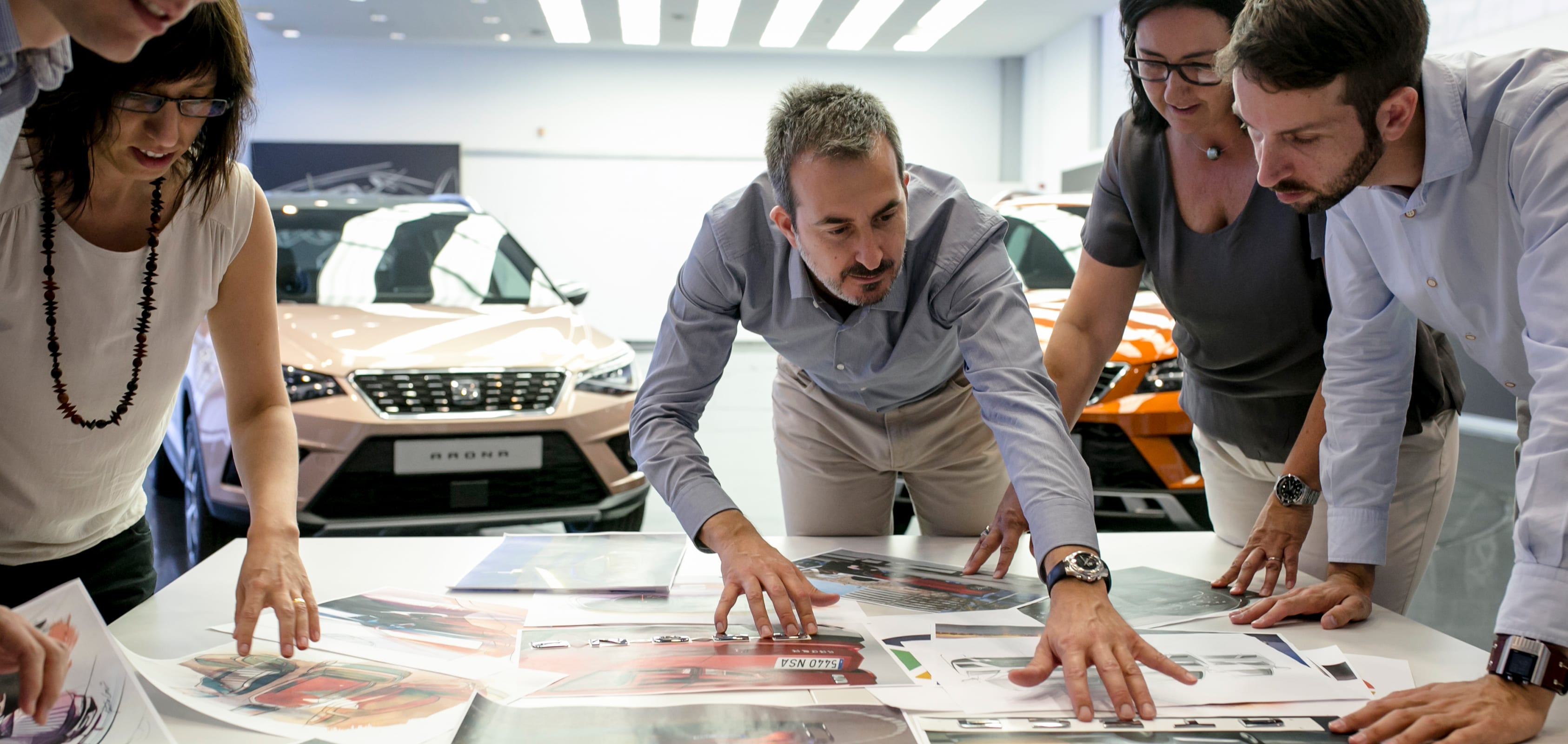 Fyra människor inspekterar fotografier av SEAT-bildesigner med SEAT Arona-modeller i bakgrunden – SEAT Human Resources